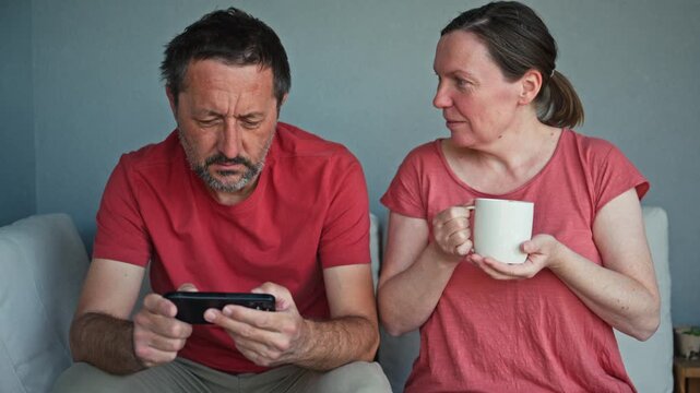 Tense middle-aged couple avoid eye contact on a couch at home; man focused on his phone while woman holds coffee mug, conveying distance, worry, disagreement and emotional tension. 4K.