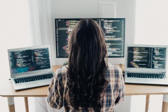 Female programmer coding on multiple laptops and a monitor in a home office, remote software developer working at a desk with code editors open on screens