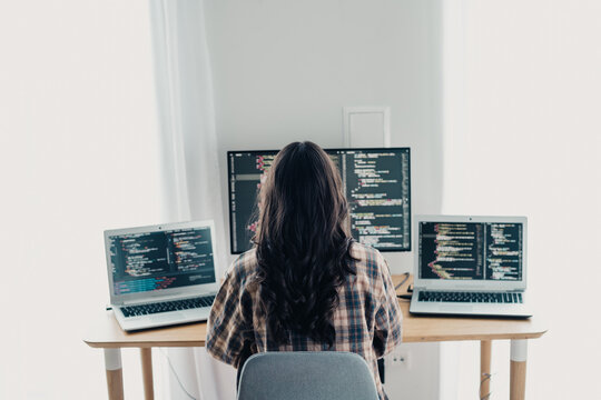 Woman programmer coding on multiple screens in a tidy home office workstation with laptops and a monitor, remote software developer focused on web development and multitasking with code