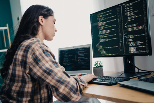 Female programmer coding at a home desk with dual monitors and laptop, focused software developer working on web code in a casual home office environment