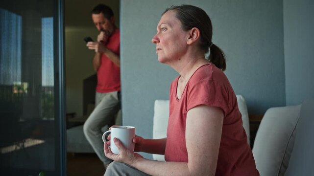 Couple at home in quiet moment, woman sitting with coffee and reflective expression while man checks phone in background, emotional distance theme. 4K.