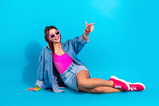 Young woman in pink swimsuit and denim jacket posing on blue background wearing sunglasses and sneakers, retro summer fashion portrait conveying fun and confident vibrant style