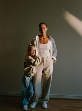 Mother and Daughter Standing Together in Matching Neutral Outfits Against Warm Wall With Natural Window Light in Studio Portrait