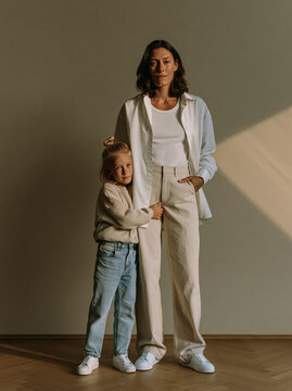 Mother and Daughter Standing Together in Matching Neutral Outfits Against Warm Wall With Natural Window Light in Studio Portrait