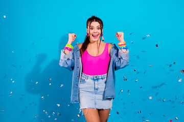 Obraz premium Excited young woman celebrating with confetti in bright pink top and denim jacket against blue background, joyful party mood and colorful accessories