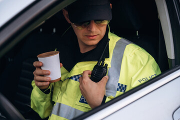 Male police officer in yellow reflective jacket holding a coffee cup and radio inside a patrol car, wearing sunglasses and focused on communication device © standret