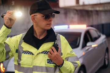 Male police officer in reflective yellow jacket and sunglasses holds flashlight while standing near patrol car with emergency lights activated in urban setting © standret