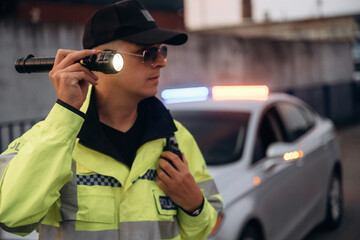 Male police officer wearing reflective yellow jacket and sunglasses holds flashlight while standing beside patrol car with emergency lights activated in urban setting © standret