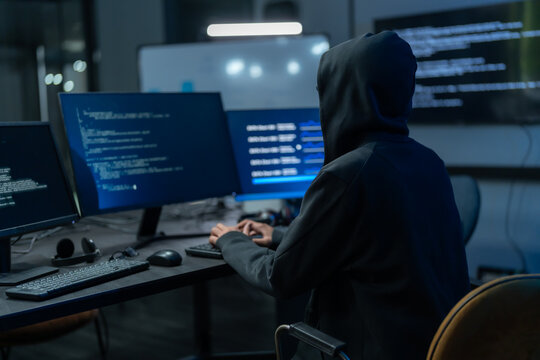 Hooded Hacker Coding On Multiple Screens In Dark Office