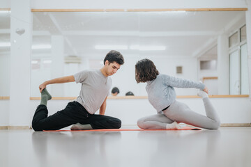 Obraz premium Two dancers perform seated partner stretches on mats in a bright studio. A male and female dancer practice yoga-like poses together beside a mirrored wall