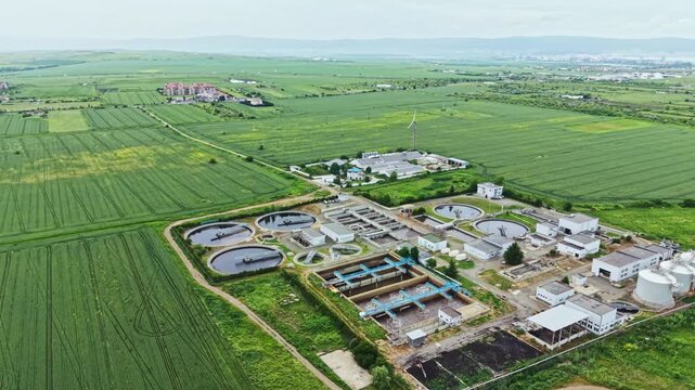 Aerial view wastewater treatment plant surrounded by green farmland and irrigation fields, circular tanks and oxidation ponds, processing buildings and access