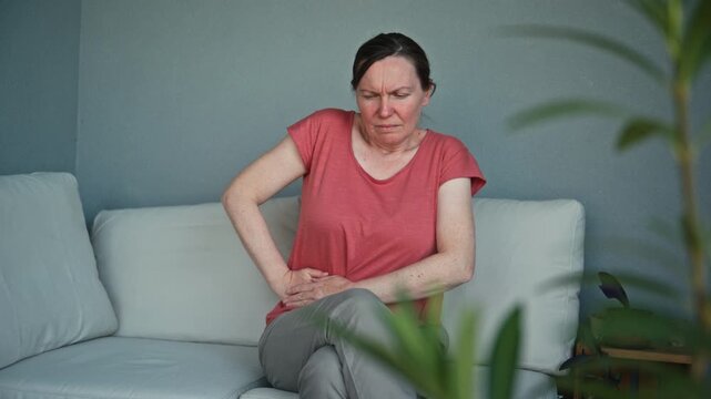 Mature woman sitting on balcony sofa holding lower right abdomen in pain. Concept of appendicitis symptoms, abdominal discomfort and health problems. 4K.