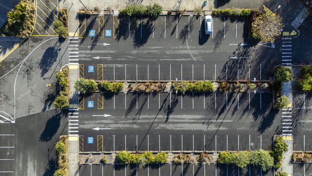 Aerial view of a parking lot with marked spaces, handicap symbols, and a single white car parked among green trees and long shadows Nelson, Nelson Region, New Zealand.