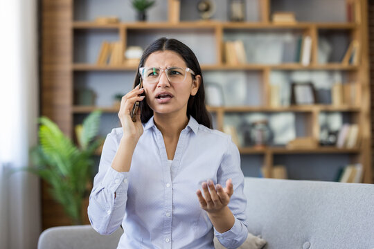 Stressed young indian woman on a couch in a home office, talking on her mobile phone and gesturing in frustration during a difficult remote work conversation
