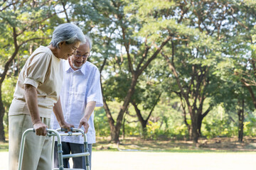 Obraz premium Senior Asian man supporting an elderly woman using a walker in the park. Happy grandfather helping his wife with a walking frame, isolated on a natural green background with copy space for text