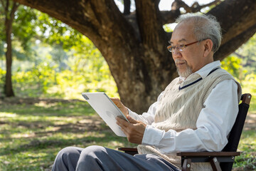 Fototapeta premium Senior Asian man using a digital tablet while sitting on a chair in the park. Elderly grandfather wearing eyeglasses looking at a screen under a tree, isolated on a natural green background