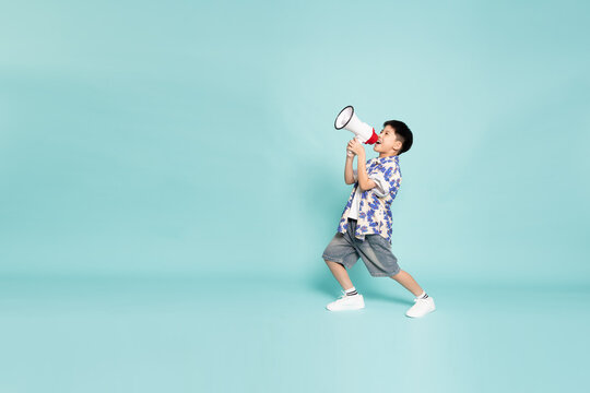 Full body of a happy Asian boy shouting into a megaphone to announce news. Cheerful young kid in a casual summer shirt calling out loud, isolated on a blue studio background with large copy space