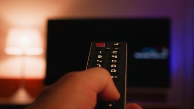 A close-up of a hand holding a TV remote control and changing channels. The TV screen is blurred in the background, creating a modern concept of home entertainment and relaxation. High quality 4k