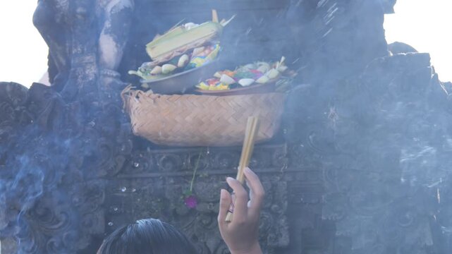 Balinese Hindu offering ceremony, with a woman placing flowers and incense on a ceremonial tray. Rising smoke creates a spiritual atmosphere during a temple ritual in Bali, Indonesia