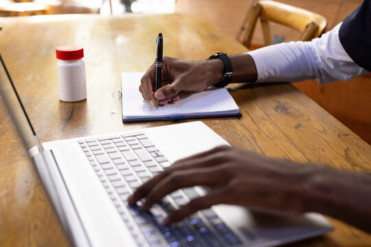 Adult African man typing and writing at wooden table with laptop, notebook, pen, wristwatch, bottle