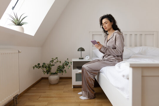 Woman sitting on side of her bed and using cellphone, checking messages and notifications in the morning in cozy bedroom