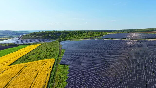 Solar panels cover a large area in the countryside. Green fields surround the panels, with patches of yellow crops present. The sky is clear and bright above the landscape.
