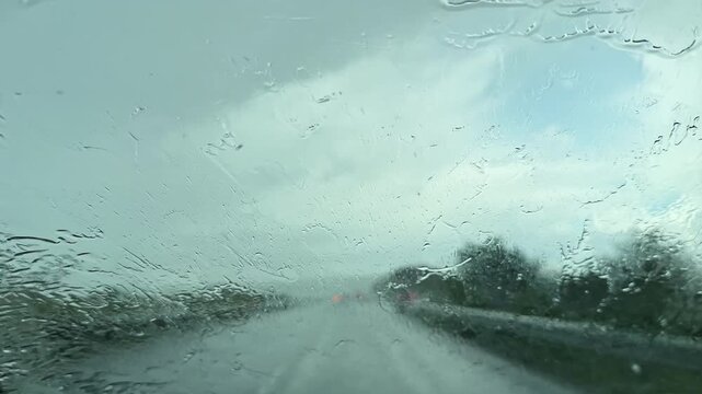 Rain is falling on a highway as a car drives. The windshield wipers work to clear droplets. Gray clouds cover the sky, and the roadway appears wet and reflective.