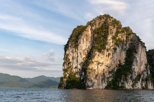 Majestic limestone cliff face, lush green jungle, and calm water under a soft blue sky with wispy clouds.