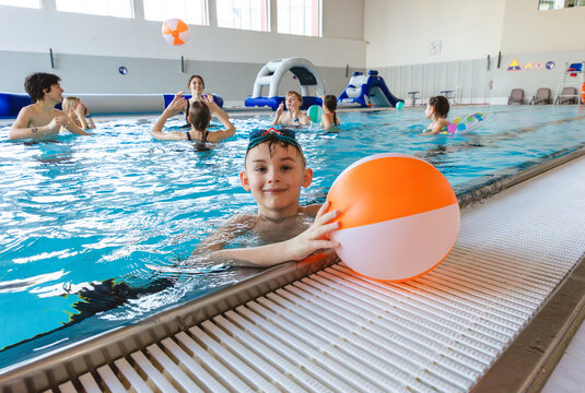 Smiling young boy with goggles leaning on the edge of an indoor swimming pool with people playing in the background
