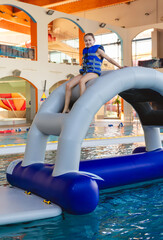 Smiling young girl wearing a life vest sitting on top of an inflatable bridge section of a water obstacle course in a public pool