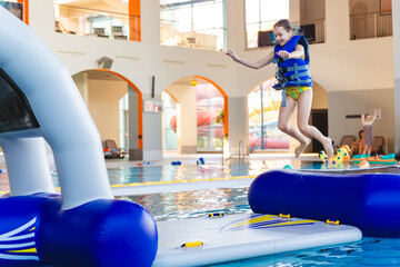 Smiling young girl wearing a life vest sitting on top of an inflatable bridge section of a water obstacle course in a public pool