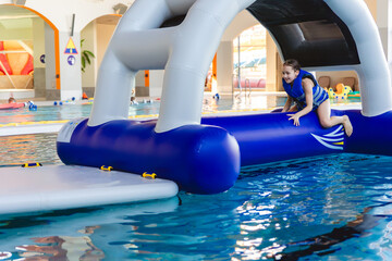 Smiling young girl wearing a life vest sitting on top of an inflatable bridge section of a water obstacle course in a public pool