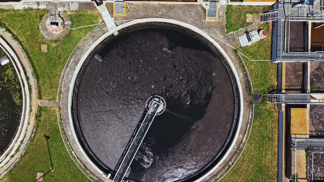 Aerial view circular clarifiers wastewater plant, drone perspective showing round settling tanks and mechanical arms skimming foamy surface, grassy buffer