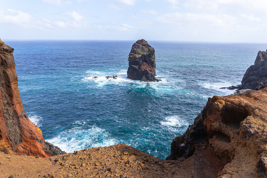 High-angle view of rugged reddish volcanic cliffs and a solitary sea stack surrounded by turquoise ocean water under a cloudy sky at Ponta de Sao Lourenco, Madeira.