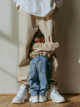 Small Child Hugging Parent Leg From Behind in Denim Jeans and White Sneakers Against Warm Beige Wall in Tender Family Moment