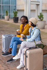 Fototapeta premium Diverse couple sitting on wooden bench outside modern entrance, holding coffee cups near suitcases