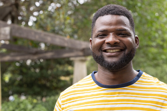 African American man standing, smiling, wearing striped yellow tee with navy collar by pergola