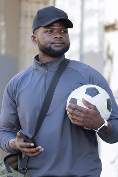 Man wearing dark baseball cap and gray jacket, holding soccer ball and checking smartphone by wall