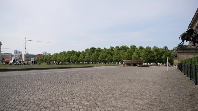 A broad cobblestone plaza opens beside leafy trees and classical museum architecture in central Berlin. This preview shows calm public space, summer light, and urban heritage scenery.