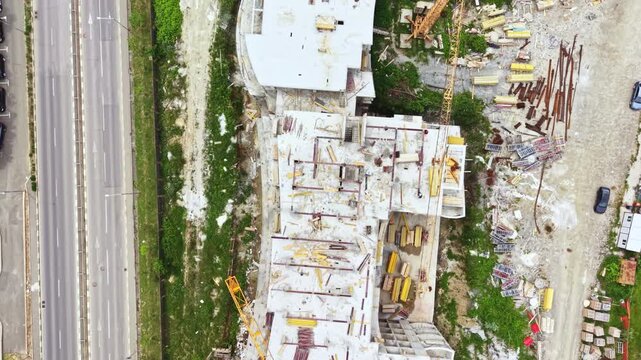 Aerial construction site beside highway, drone view of demolition and rubble with concrete shells and heavy machinery, traffic flowing on adjacent lanes, grassy