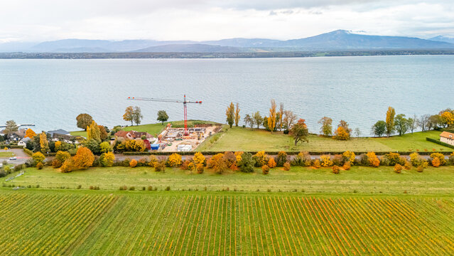 Aerial view of a construction site with a red crane, autumn trees, and vineyards on the shores of Lake Geneva in Crans VD, Vaud, Switzerland.