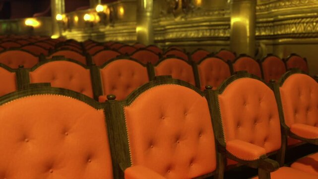 Empty red velvet seats under chandelier, gold balconies shimmer with dim amber lighting, rows of tufted armchairs create intimate vintage atmosphere, plush