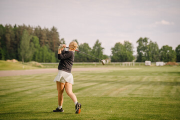 Obraz premium Female golfer in black sweater and white skirt swings golf club on green course with trees and open field in the background under a clear sky