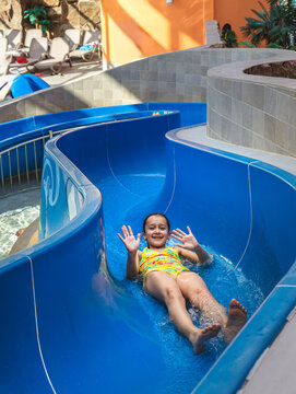 Happy young girl waving hands while sliding down a blue water slide at an indoor water park