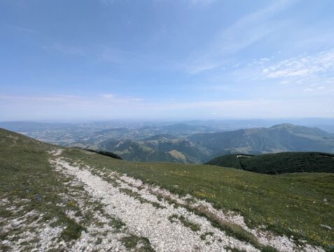 Sentiero roccioso su pendio erboso che scende verso una valle ampia con montagne e pianure lontane, sotto un cielo limpido con poche nuvole