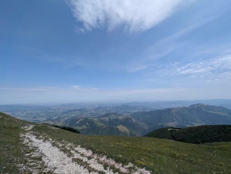 Sentiero roccioso su pendio erboso che scende verso una valle ampia con montagne e pianure lontane, sotto un cielo limpido con poche nuvole