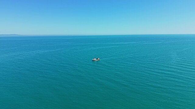 A boat is seen fishing on a vast blue ocean. The water stretches out far with smooth waves. The sky is clear and bright. It is mid-day and the sun is shining.