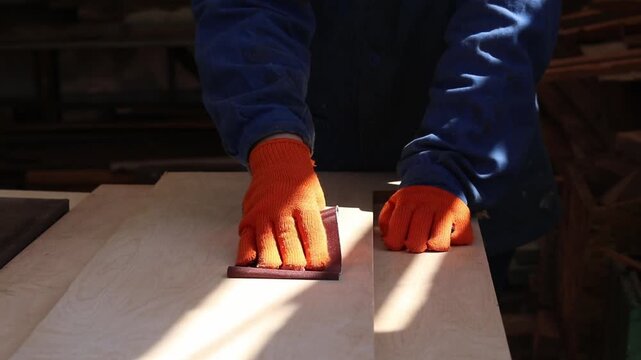 A hand in an orange glove sands a wooden surface with sandpaper. The master processes a wooden board manually using sandpaper. The process of preparing wood for painting or varnishing in a workshop