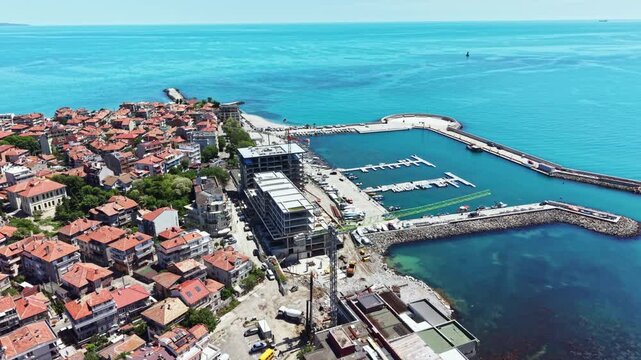 A seaside town with many buildings and homes along the shore. There is a marina with boats docked. Construction is ongoing near the waterfront under a clear blue sky.