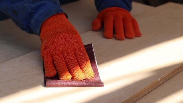 An unrecognizable person in blue work clothes and protective gloves is working on the processing of a wooden board. The process of sanding the surface of a board using sandpaper. Handicraft, hobby